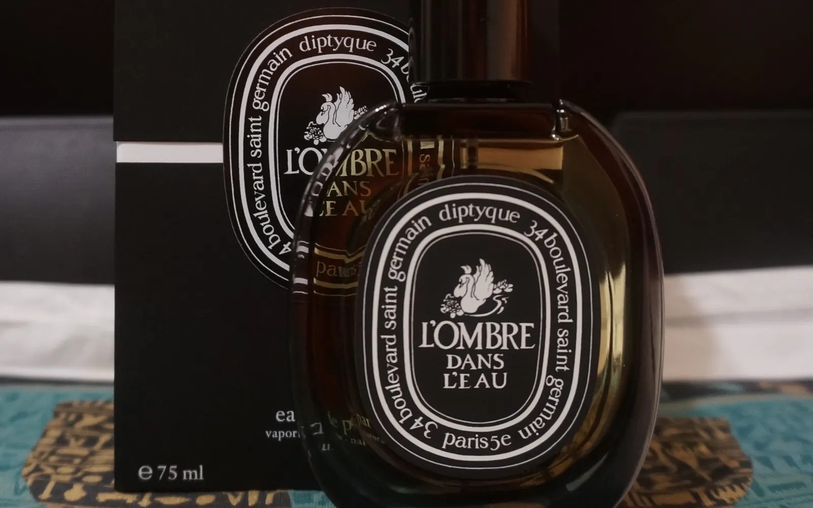 A Diptyque L’Ombre dans l’Eau bottle in front of its carton, which is printed with ℮ 75 ml